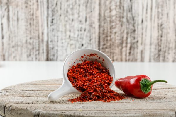 Chili powder with red pepper and wooden piece in a white scoop on white and wooden grunge background, side view.